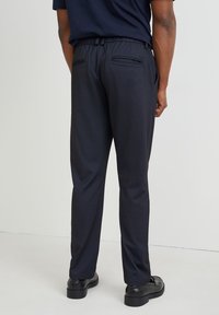 Pantalon marine en tissu texturé, taille élastique, deux poches arrière et jambes fuselées, accompagné de chaussures noires.