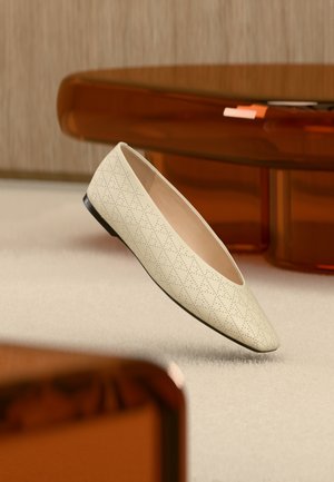 Chaussure plate beige avec des perforations géométriques flottant au-dessus d'un tapis clair dans une pièce avec des meubles de couleur ambre.
