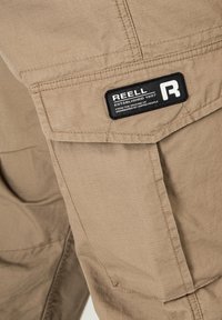 Beige Cargo-Hosen aus strukturiertem Stoff, mit einer Seitentasche und einem schwarzen Etikett mit der Aufschrift "REELL" sowie markanten Nähten.