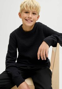 Must have mustrisümpa, millel on soonikultuuriga ümar kaelus ja varrukate lõpp. Tekstuur näib pehme, kombineeritud mustade pükstega. Taustal on hele puidust tool.