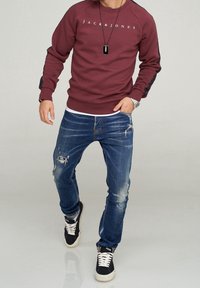 Vinröd sweatshirt med logotyp på framsidan, svarta detaljer på ärmarna, kombinerad med slitna blå jeans och svarta sneakers med vita sulor.