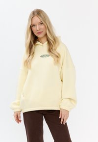 Felpa gialla oversize con cappuccio e cordino, caratterizzata da un logo ricamato verde sul petto, texture liscia e maniche con polsino.