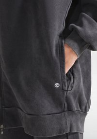 Felpa grigia scura con zip anteriore, polsini a costine e tasche laterali. Presenta un piccolo logo sul davanti. Tessuto morbido e di consistenza.