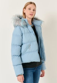 Giacca puffer azzurro chiaro con cappuccio in pelliccia sintetica, tasche con zip e design cropped. Presenta una texture liscia e lucida e polsini elasticizzati.