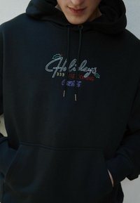 Melnā kapucīna krekls ar daudzkrāsainu izšūtu tekstu "Brīvdienas nāk", ar dekoratīviem akcentiem. Izstrādāts ar kangaroo kabatu un aukliņām.