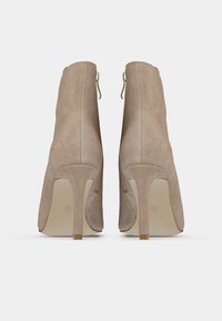 Botines de ante beige con un tacón alto delgado, que cuentan con una cremallera en la parte trasera y una textura suave. Diseño elegante y ajustado.