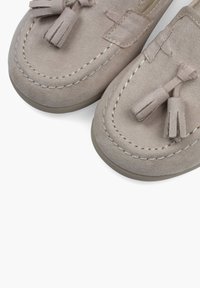 Beige mokkasiner i semsket skinn med sømdetaljer og dusker på toppen, med avrundede tåer og gummisåler.