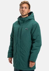 Zelená zateplená parka s kapucňou, predným zipsom a bočnými vreckami. Hladká textúra a logo na ľavej strane hrudníka. Obsahuje nastaviteľné prvky.