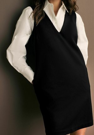 Femme portant une robe noire sans manches à col en V par-dessus une chemise blanche à manches longues avec boutons, debout devant un fond neutre.