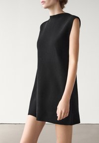 Femme portant une robe noire sans manches, de longueur au genou, coupe trapèze, debout devant un fond blanc et gris uni.