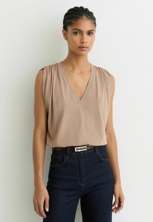 Naine punutud juustega, kes kannab varrukateta beežikat V-kaelusega topi, mis on tumesiniste teksade sisse toppitud ja millel on õhuke pruun vöö, seisab valge seina taustal.