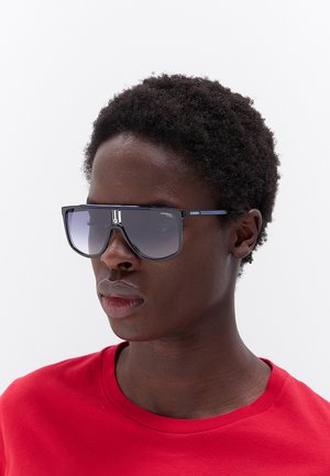Persona con capelli corti e ricci che indossa grandi occhiali da sole scuri e una camicia rossa su uno sfondo chiaro uniforme.