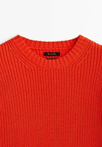 Pull en tricot rouge épais avec un col rond, des poignets et un ourlet côtelés. Présente un motif texturé. L'étiquette affiche le nom de la marque.