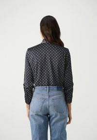 Blusa nera a pois con maniche lunghe e colletto, abbinata a jeans azzurri a vita alta. Texture liscia, motivo classico.