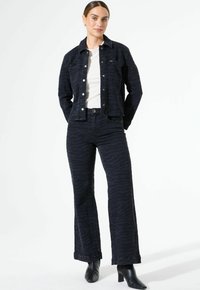 Veste en denim foncé et pantalons évasés avec un motif subtil de rayures zèbre. La veste est dotée de boutons-pression ; elle est associée à un haut clair et à des bottes noires.