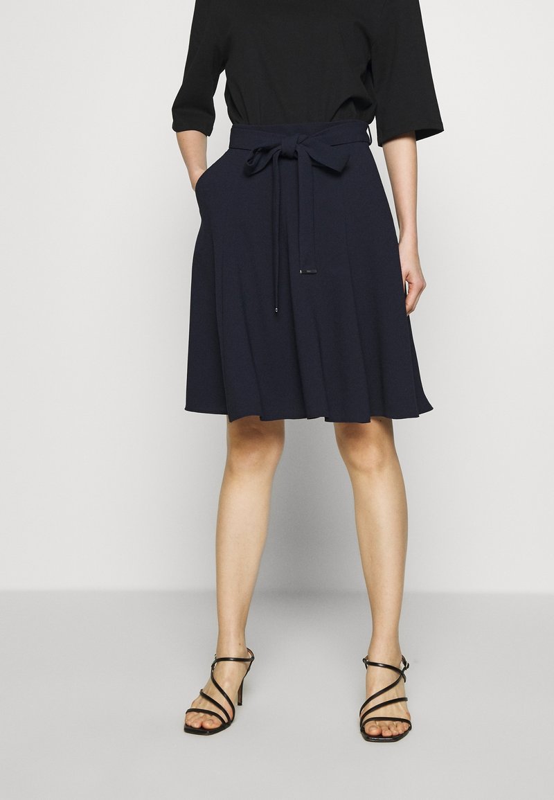 Femme portant une jupe bleu marine longueur genou avec une ceinture nouée, un haut noir à manches coudées et des sandales noires à talons hauts avec des lanières.