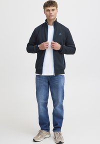 Mørkeblå lynlåstrøje med høj krave, blå lynlåsdetaljer og et logo på brystet, kombineret med blå jeans og grå sneakers.