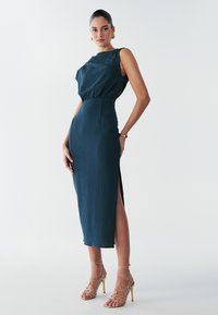 Robe midi bleu sarcelle avec une finition texturée, sans manches, taille ceinturée et fente sur le côté. Associée à des sandales à talons hauts dorées à lanières.