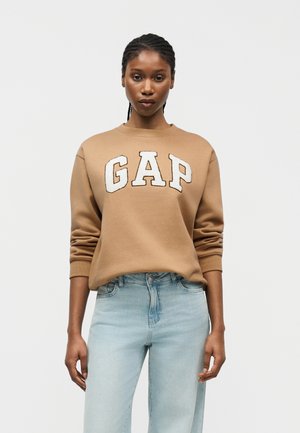 Frau mit geflochtenem Haar, die einen hellbraunen GAP-Sweatshirt und hellblaue Jeans trägt, stehend vor einem schlichten weißen Hintergrund.