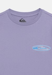 Μπλουζάκι σε χρώμα λεβάντας με στρογγυλή λαιμόκοψη, με μικρό μπλε και λευκό οβάλ λογότυπο κύματος στο αριστερό στήθος και μαύρο λογότυπο κύματος στην εσωτερική ετικέτα στον λαιμό.