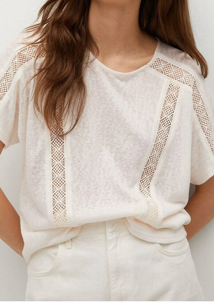 Haut blanc à manches courtes avec des bandes en dentelle à motifs sur les épaules et l’avant, porté rentré dans un pantalon blanc taille haute.