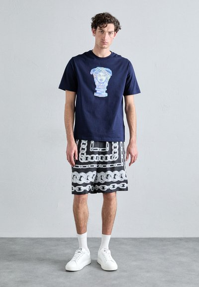 T-shirt en coton bleu marine avec un graphique multicolore sur la poitrine, associé à un short noir arbordant un motif de chaîne blanche. Complété par des baskets blanches.