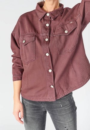 Kvinde iført en løstsiddende mauve skjortejakke med knapper og klaplommer, kombineret med grå slidte jeans, med den ene arm hævet.