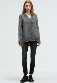 Eine Frau trägt ein lockeres graues Hemd mit Knöpfen und schwarzem Schriftzug, schlanke schwarze Jeans und schwarze Turnschuhe und steht frontal.