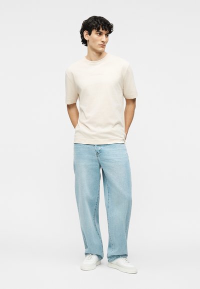 Νέος άνδρας με σγουρά μαλλιά, φορώντας ένα μπεζ πουκάμισο με κοντά μανίκια, ανοιχτό μπλε τζιν παντελόνι φαρδιάς γραμμής και λευκά αθλητικά παπούτσια, στέκεται με τα χέρια στις τσέπες.