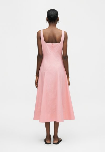 Γυναίκα με τα μαλλιά σε κότσο φορά αμάνικο φόρεμα μέτριου μήκους σε ανοιχτό ροζ χρώμα με τετράγωνο λαιμόκοψη στην πλάτη και μαύρα επίπεδα σανδάλια, στέκεται μπροστά σε απλό φόντο.