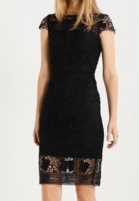 Musta pitsimekko, jossa on lyhyet hihat, aaltoilevat reunat ja tiukka siluetti. Huomattavat kukkakuviot ja läpikuultava alaosa.