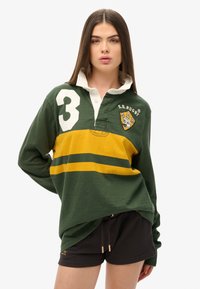 T-shirt de style rugby vert avec une rayure jaune, doté d'un col blanc, du numéro 3 et d'un écusson de logo. Le matériau semble doux et texturé.