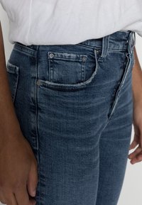 Jeans de mezclilla azul con un corte ajustado, que presentan bolsillos frontales, remaches de metal y un dobladillo deshilachado a lo largo de la cinturilla. Tejido texturizado.