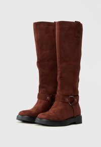 Botas de ante marrón hasta la rodilla con una suela negra plana, que presentan un acabado suave, una correa en el tobillo con un anillo metálico y un diseño simple.