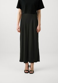 Top scurt negru, fustă maxi neagră, fluidă, dintr-un material neted, asortată cu sandale negre cu accente aurii. Design simplu, siluetă elegantă.
