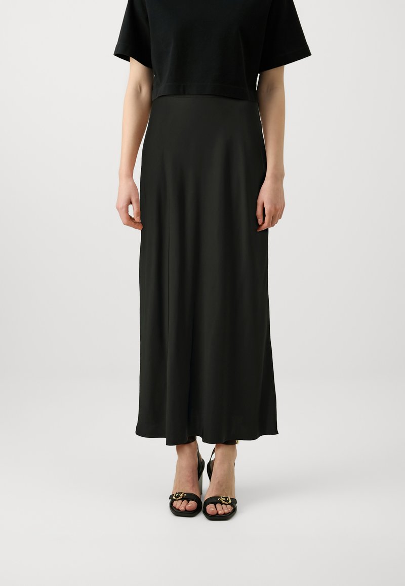 Top scurt negru, fustă maxi neagră, fluidă, dintr-un material neted, asortată cu sandale negre cu accente aurii. Design simplu, siluetă elegantă.