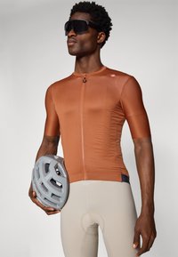 Maillot de cyclisme marron à manches courtes et zip sur le devant, associé à un short beige. Le modèle tient un casque gris avec des trous de ventilation.