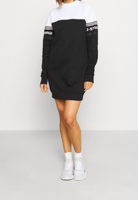 Kvinna som bär en svart och vit långärmad casual klänning med varumärkestext på ärmarna, i kombination med vita knästrumpor och vita sneakers.