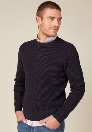 Pull en maille bleu marine avec un motif texturé, col rond, poignets et ourlet côtelés, superposé à une chemise claire à col.