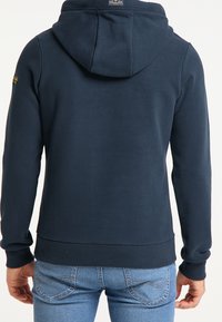 Man i en marinblå huvtröja och ljusblå jeans, stående med avslappnade händer vid sidorna, med ryggen vänd mot betraktaren.