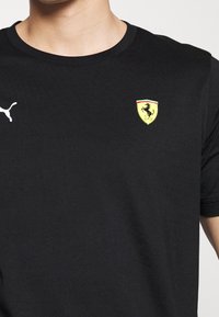 Černé bavlněné tričko s bílým logem Puma na levé části hrudi a žluto-červeným emblémem Ferrari se skákajícím hřebcem.