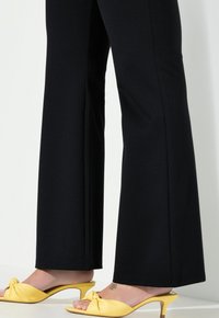 Pantalons larges noirs avec une texture lisse, associés à des mules à talons jaunes présentant un détail en nœud sur le dessus.