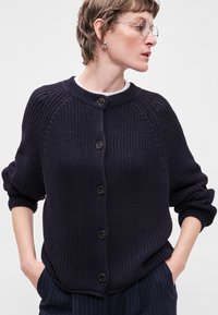 Ναυτικό μπλε πλεκτή ζακέτα με ραγλανά μανίκια, κλείσιμο με κουμπιά μπροστά, υφασμάτινη επιφάνεια και ριγωτές μανσέτες. Φορεμένη πάνω από ένα λευκό πουκάμισο.