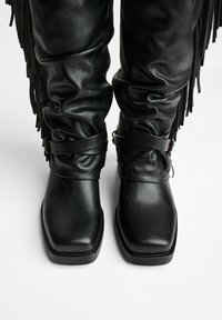 Black leather knee-high boots with a slouchy design, square toe, and decorative fringe. Features two black straps around the calf.