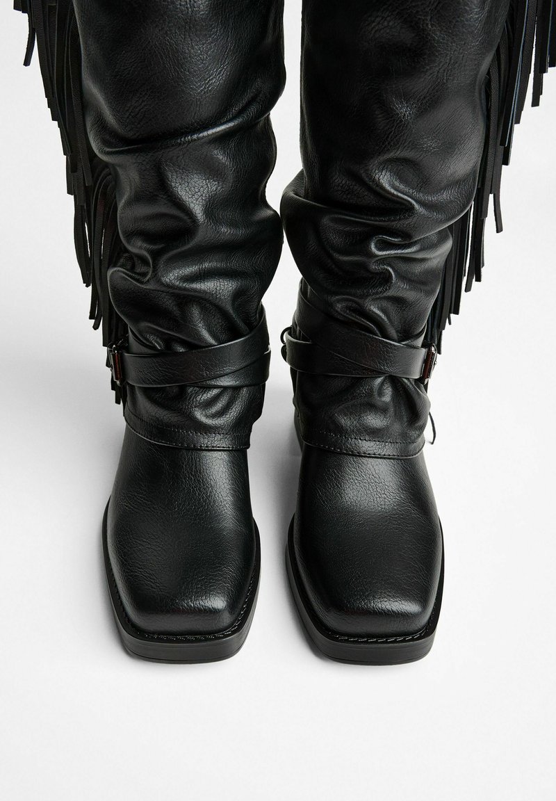 Black leather knee-high boots with a slouchy design, square toe, and decorative fringe. Features two black straps around the calf.