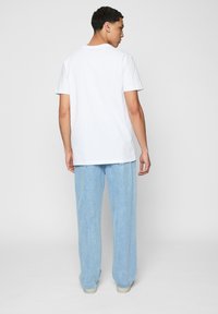Ung mand står med ryggen til, iført en simpel hvid T-shirt med kort ærme og lyseblå vidde jeans mod en ensfarvet baggrund.