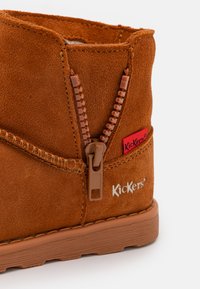 Botín de ante marrón con cremallera lateral, detalle de costura y suela de goma. Presenta un pequeño logo de marca rojo en el lateral.