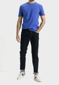 Camiseta de algodón azul de manga corta con un bolsillo en el pecho, combinada con jeans de mezclilla oscura y zapatillas blancas con acentos marrones y puños remangados.