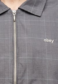 Γκρι καρό μπουφάν με μπροστινό μεταλλικό φερμουάρ και το κέντημα "obey" στο δεξί στήθος, που δείχνει μέρος του γιακά και του λαιμού.