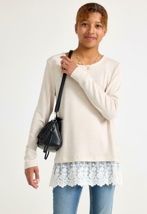 Ung kvinde iført en beige langærmet top med blonder, blå jeans, en halskæde og bærer en sort håndtaske på skulderen.